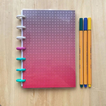 A Tiny Notes Notebook with a gradient red to purple cover and dotted pattern on the front is placed on a wooden surface. The notebook, MAMBI Happy Planner compatible, has multicolored rings on the left side. Next to the notebook are three striped pens in yellow, orange, and blue.