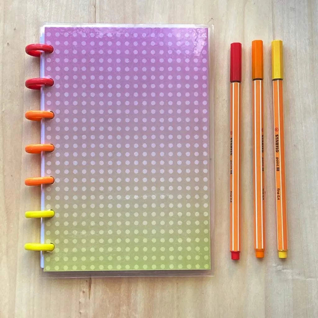 A Tiny Notes Notebook with a gradient cover transitioning from purple at the top to green at the bottom, with white polka dots, lies on a wooden surface. This refillable discbound notebook features red, orange, and yellow rings. Three matching pens in red, orange, and yellow are beside it.