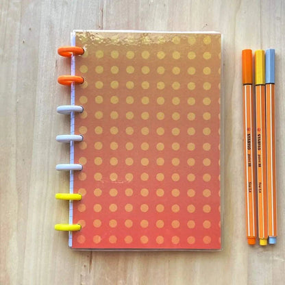 A Tiny Notes Notebook with a gradient orange and yellow polka dot cover and multicolored rings is placed on a wooden surface. Beside it, three pens in orange, yellow, and gray are lined up neatly. This MAMBI Happy Planner compatible notebook is also fountain pen friendly.