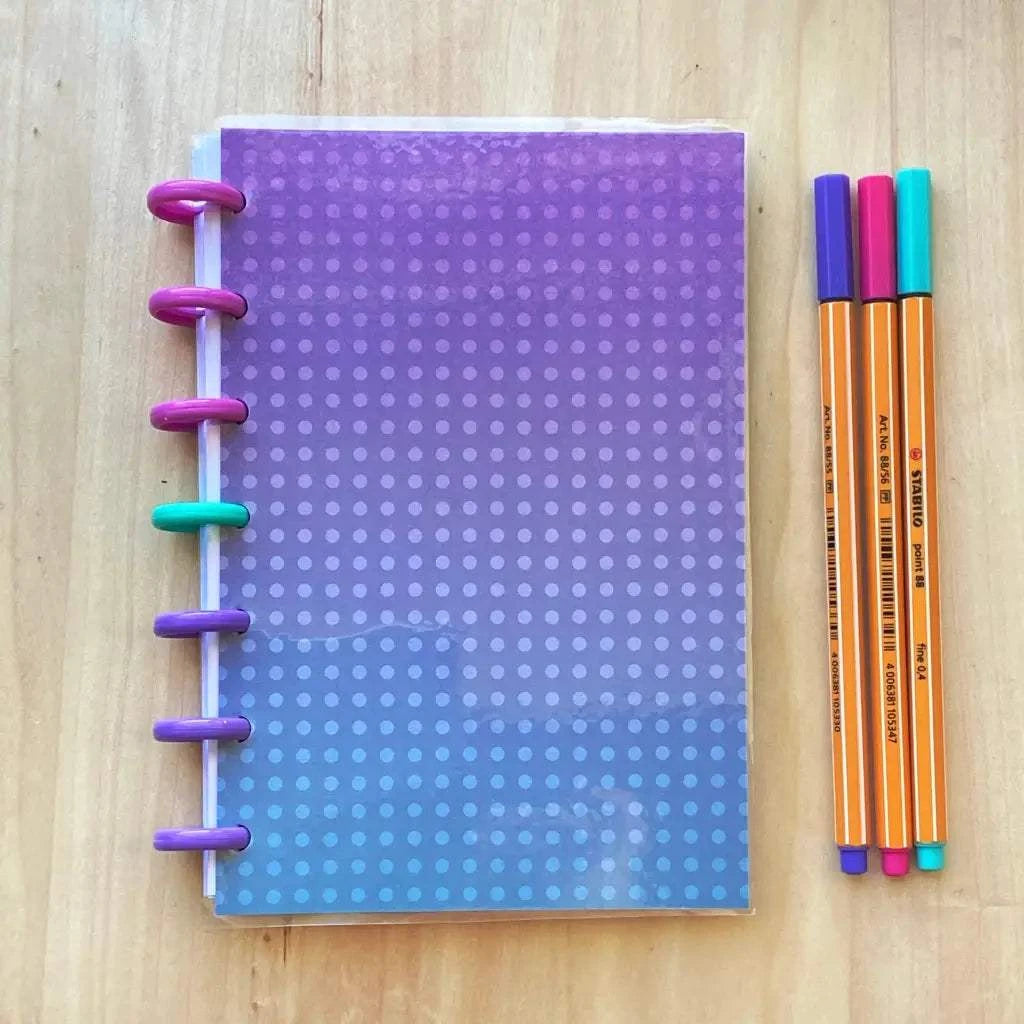 A colorful, fountain pen friendly Tiny Notes Notebook with a purple to blue gradient cover and a polka dot pattern lies on a wooden surface. The refillable discbound notebook, MAMBI Happy Planner compatible, boasts multicolored disc bindings on the left side. Next to it are three markers in orange, purple, and blue.