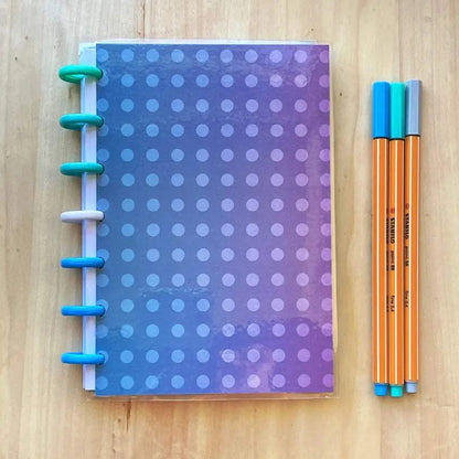 A polka dot notebook with a gradient blue and purple cover lies on a wooden surface next to three orange fineliner pens, each with blue caps. This refillable Tiny Notes Notebook has large colorful binding discs on the left side and is MAMBI Happy Planner compatible.