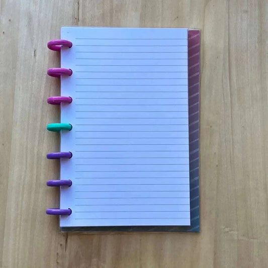 A Tiny Notes Notebook with colorful disc rings is placed on a wooden surface. The rings are in gradient colors, including pink, purple, turquoise, and green. The notebook's white pages have faint blue lines, and the translucent cover shows a subtle pattern. It's also MAMBI Happy Planner compatible.