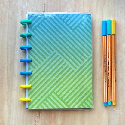 A Tiny Notes Notebook with a blue and green geometric striped cover lies on a wooden surface, bound with multicolored rings. To the right of the notebook, three fine tip pens in blue, orange, and yellow are arranged neatly. This MAMBI Happy Planner compatible accessory is also fountain pen friendly.