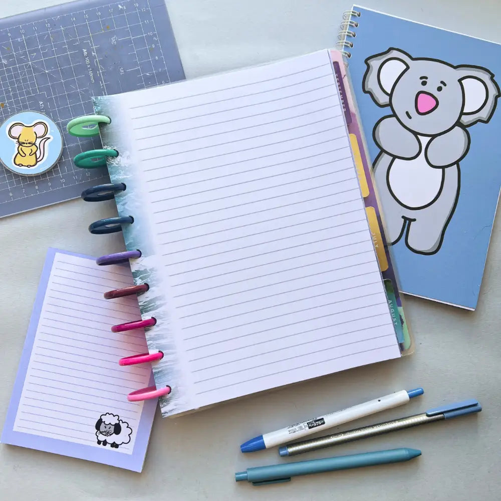 Open notebook with colorful tabs and pens on rings, surrounded by a small lined Water Themed Notepaper, a notebook featuring a cartoon koala, and various pens. Nearby is a blue cutting mat with a sticker of a squirrel. The overall setting suggests journal creation in a creative workspace.