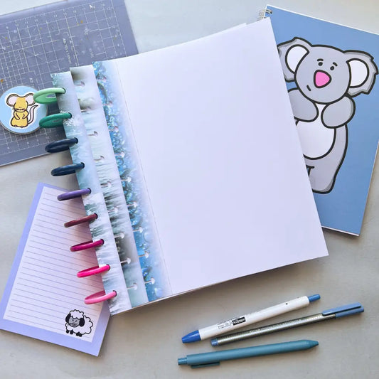 A neatly arranged desk featuring an open notebook with colored tabs, a closed koala-covered journal creation, a sheet of Water Themed Notepaper with a sheep illustration, pens, and a pencil. There is also a small square card with a squirrel sticker on it.