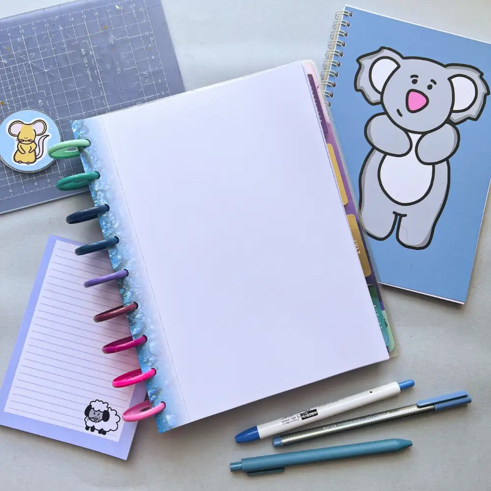 A flat lay of assorted stationery includes an open notebook with colorful rings on the spine, blank pages, two pens, a Water Themed Notepaper, and a lined notepad with a sheep illustration. A squirrel sticker is also visible on a nearby clear clipboard. Perfect for journal creation or planner note paper enthusiasts.