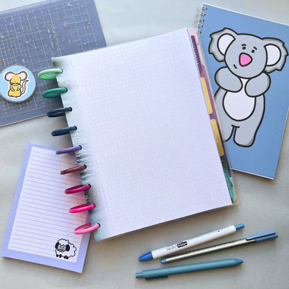 A flat lay photo of stationery items, including an open dotted spiral notebook with colorful pens clipped to the spine, a small notepad with a sheep illustration, two pens, a sticker sheet with a mouse illustration, and Water Themed Notepaper perfect for journal creation.