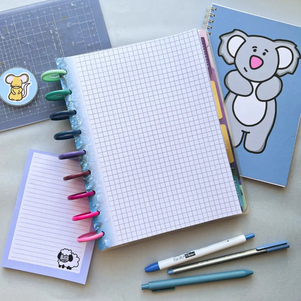 A flat lay image of a study setup includes an open graph paper notebook with colorful tabs, a smaller lined notepad with a sheep illustration, Water Themed Notepaper in a blue notebook with a koala cover, two pens, and a sticker sheet featuring squirrel stickers—a perfect scene for journal creation.