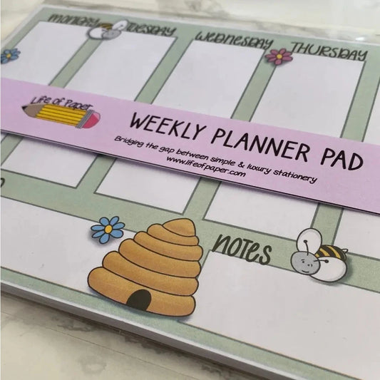 Image of an A5 size Weekly Plan Desk Memo Pad featuring sections for each day of the week, with cute illustrations of bees, flowers, and a beehive. Crafted with 120gsm paper, the planner is wrapped with a purple band that reads "Weekly Planner Pad" and includes the website "lifeofpaper.com.
