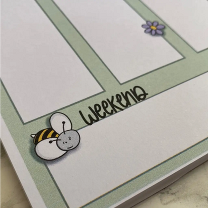 A close-up of a Weekly Plan Desk Memo Pad page with green-bordered sections on A5 size 120gsm paper. The bottom section features the word "weekend" in playful, black cursive. An illustration of a bumblebee decorates the corner, and a small purple flower is in the top right corner.