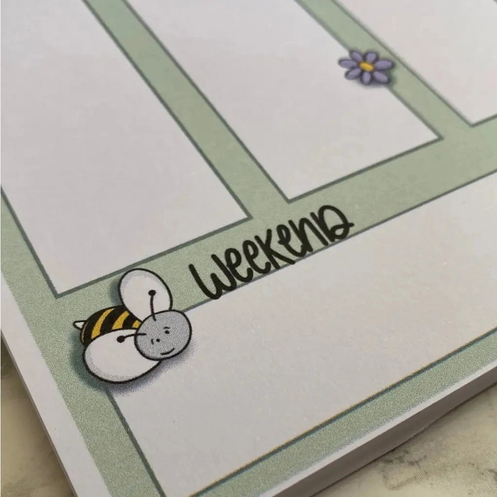 A close-up of a Weekly Plan Desk Memo Pad page with outlined boxes for writing. One section is labeled "Weekend" with a cute bee illustration in the corner. Another corner has a small purple flower illustration. The A5 size page, made from 120gsm paper, has a simple and playful design.