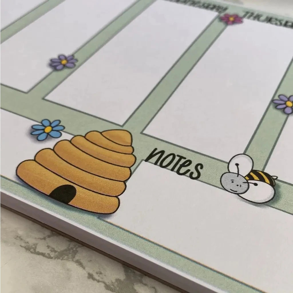 A close-up of a Weekly Plan Desk Memo Pad with the columns labeled Monday to Thursday in the background. In the foreground, the "Notes" section features an illustration of a beehive, a bee, and some small flowers. The A5 size planner, made with 120gsm paper, is resting on a marble surface.