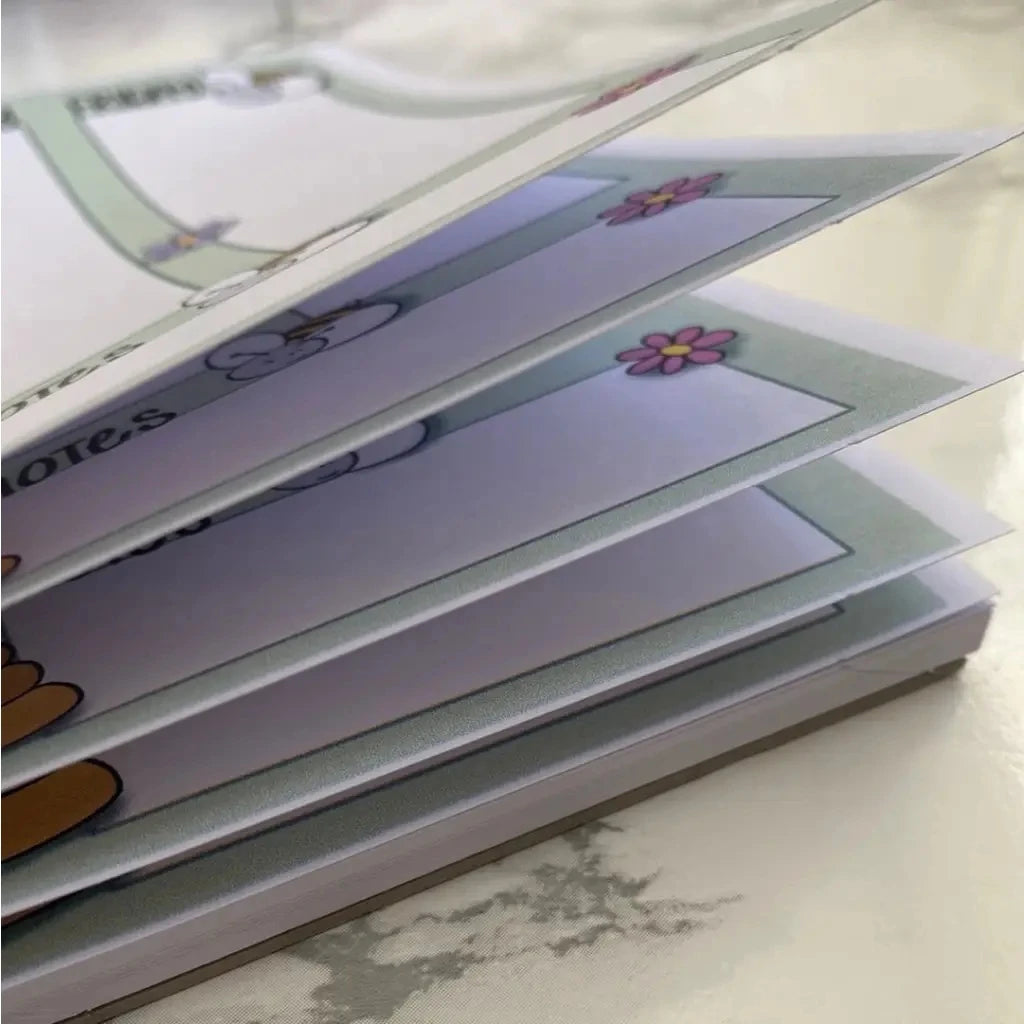 A close-up view of a partially open Weekly Plan Desk Memo Pad with illustrated pages featuring cartoon-style flowers and drawings on 120gsm paper. The images and decorations on the pages are visible, indicating that the memo pad contains playful and colorful art.