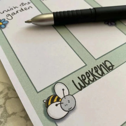 A close-up of a Weekly Plan Desk Memo Pad page with a black pen resting on top. The A5 size page, made from 120gsm paper, features a small, cute bee illustration and the word "Weekend" in decorative font. A part of a handwritten task, "Finish the garden," is visible in one section.