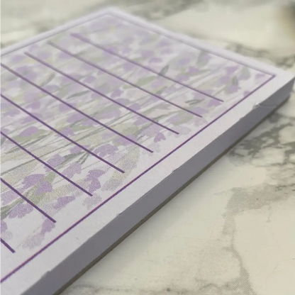 A close-up of a section of a Wisteria Memo Pad with faint purple wildflower illustrations. The notepad, ideal as a memo pad, has lined sections for writing, and the edges of the pages show a slight curl. The background has a marble-like pattern in shades of grey and white.