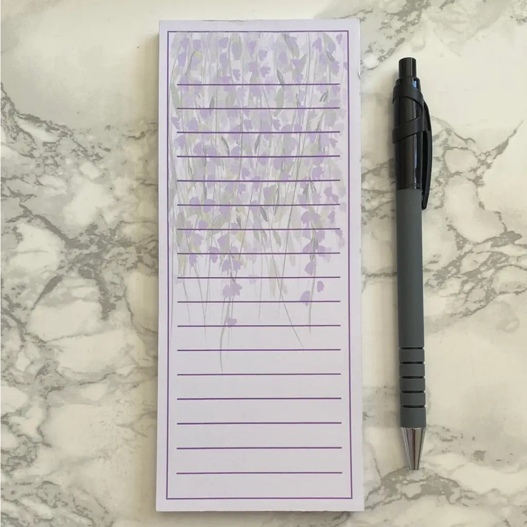 A Wisteria Memo Pad featuring lined paper with a purple floral design at the top, placed on a marble surface next to a gray pen with a black clicker top. Perfect for jotting down your to-do list or shopping list.
