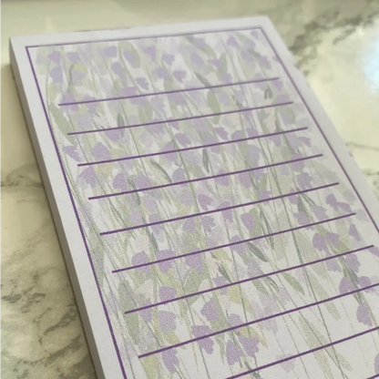 A close-up photo of a Wisteria Memo Pad with purple flowers and green stems as a background. Featuring horizontal purple lines for writing your shopping list or to do list, the notepad is placed on a surface with a light, marbled pattern.