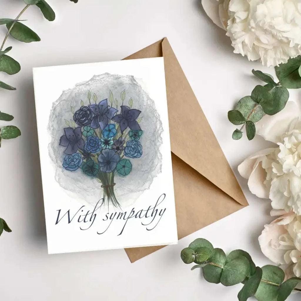 The "With Sympathy Greeting Card" showcases an illustration of blue and purple flowers on its cover. It is accompanied by a biodegradable brown envelope and features the words "With sympathy." The bouquet is beautifully surrounded by white flowers and green eucalyptus leaves.