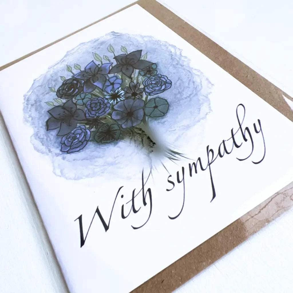 The "With Sympathy Greeting Card" features a bouquet of blue and purple flowers with green leaves at the center. Below the floral arrangement, the words "With sympathy" are elegantly written in cursive handwriting. The card comes with a biodegradable envelope for an eco-friendly touch.