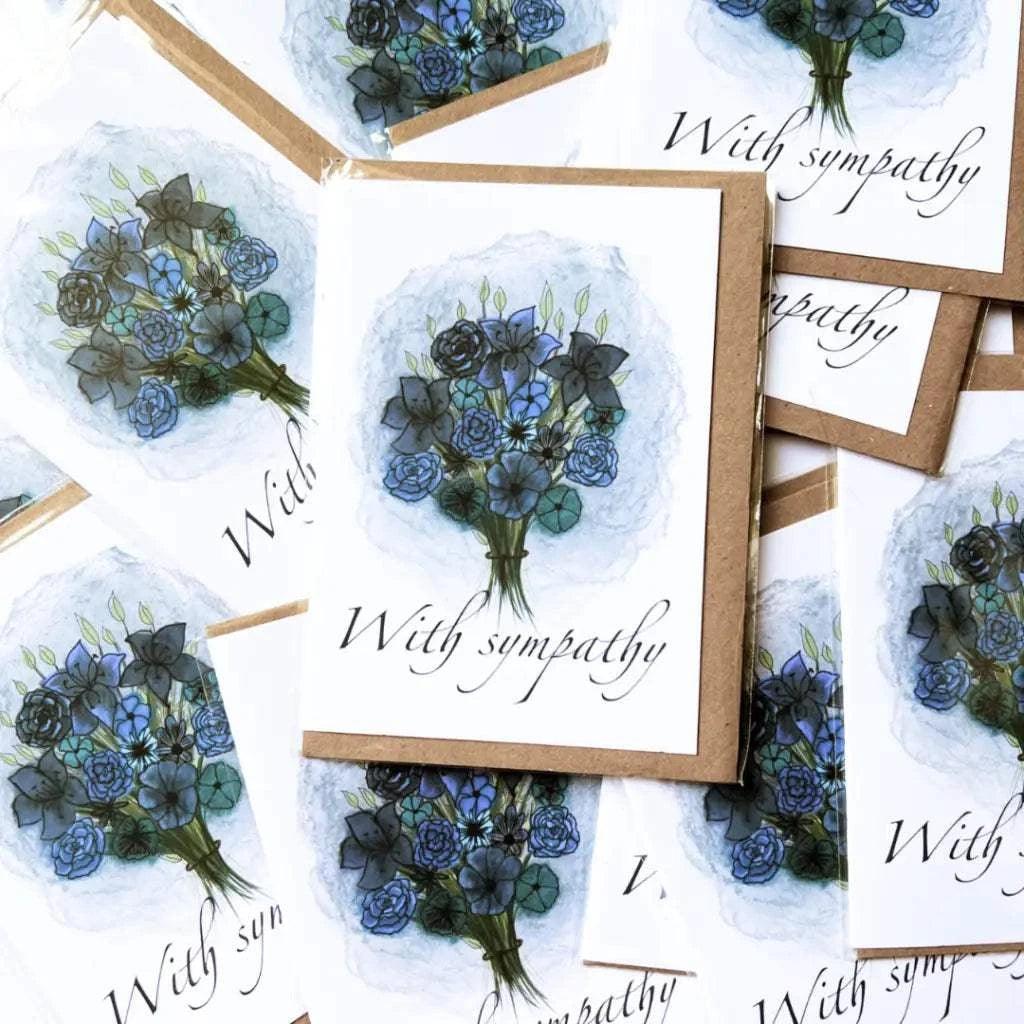 The With Sympathy Greeting Card collection features cards with an illustration of a floral bouquet in blue and black on the front, each bearing the message "With sympathy" in elegant script. The cards are arranged to partially overlap each other, presenting a variety of angles, and come with biodegradable envelopes.